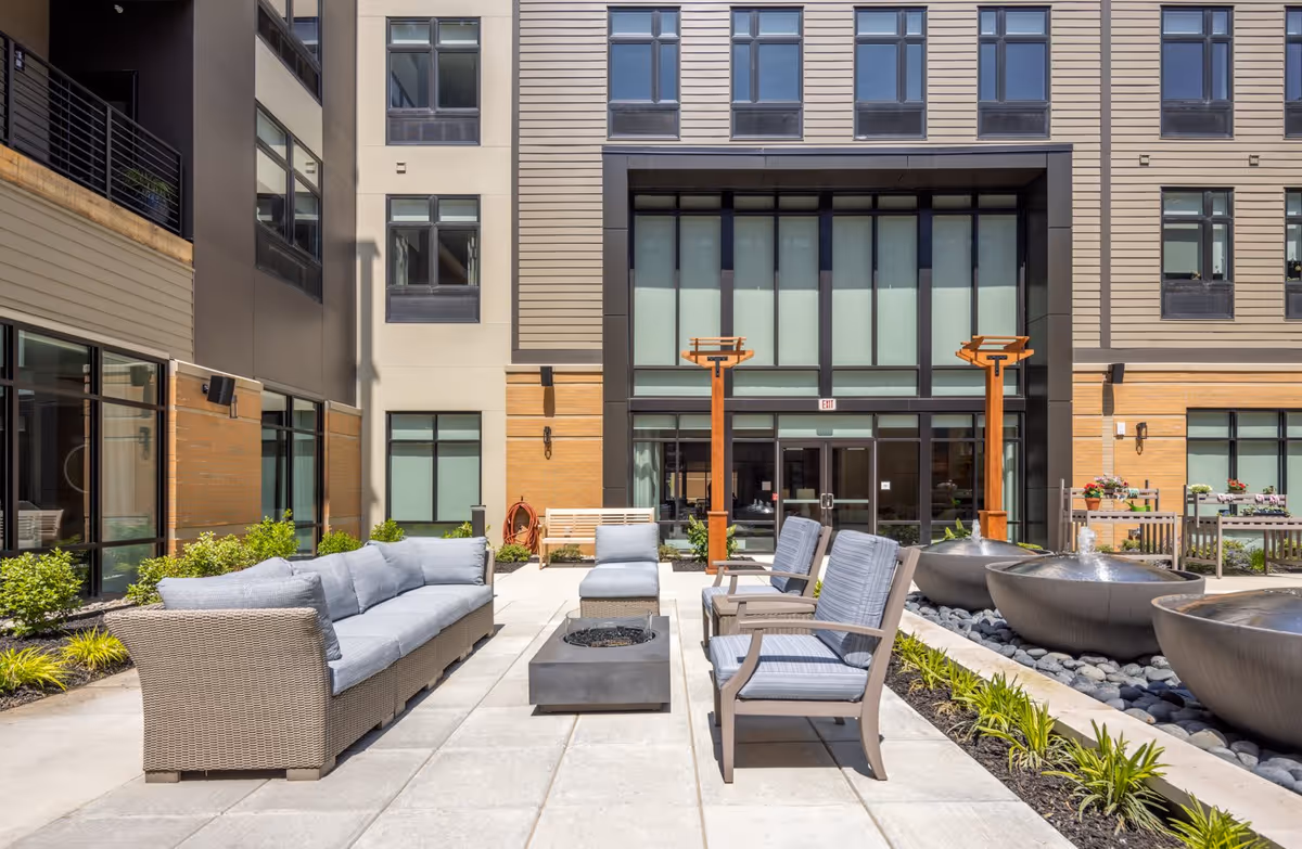 Outdoor courtyard area at The Summit of Blue Ash featuring cushioned wicker sofa, cushioned chairs, a fire pit, three large water fountains, and landscaped plants, surrounded by a multi-story building with large windows.