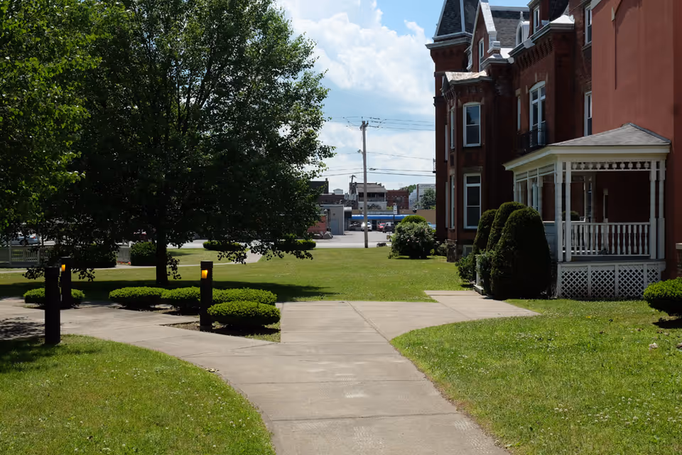 A paved walkway leading through a green lawn with trimmed bushes and trees, adjacent to a red brick building with white porch railings under a partly cloudy sky.