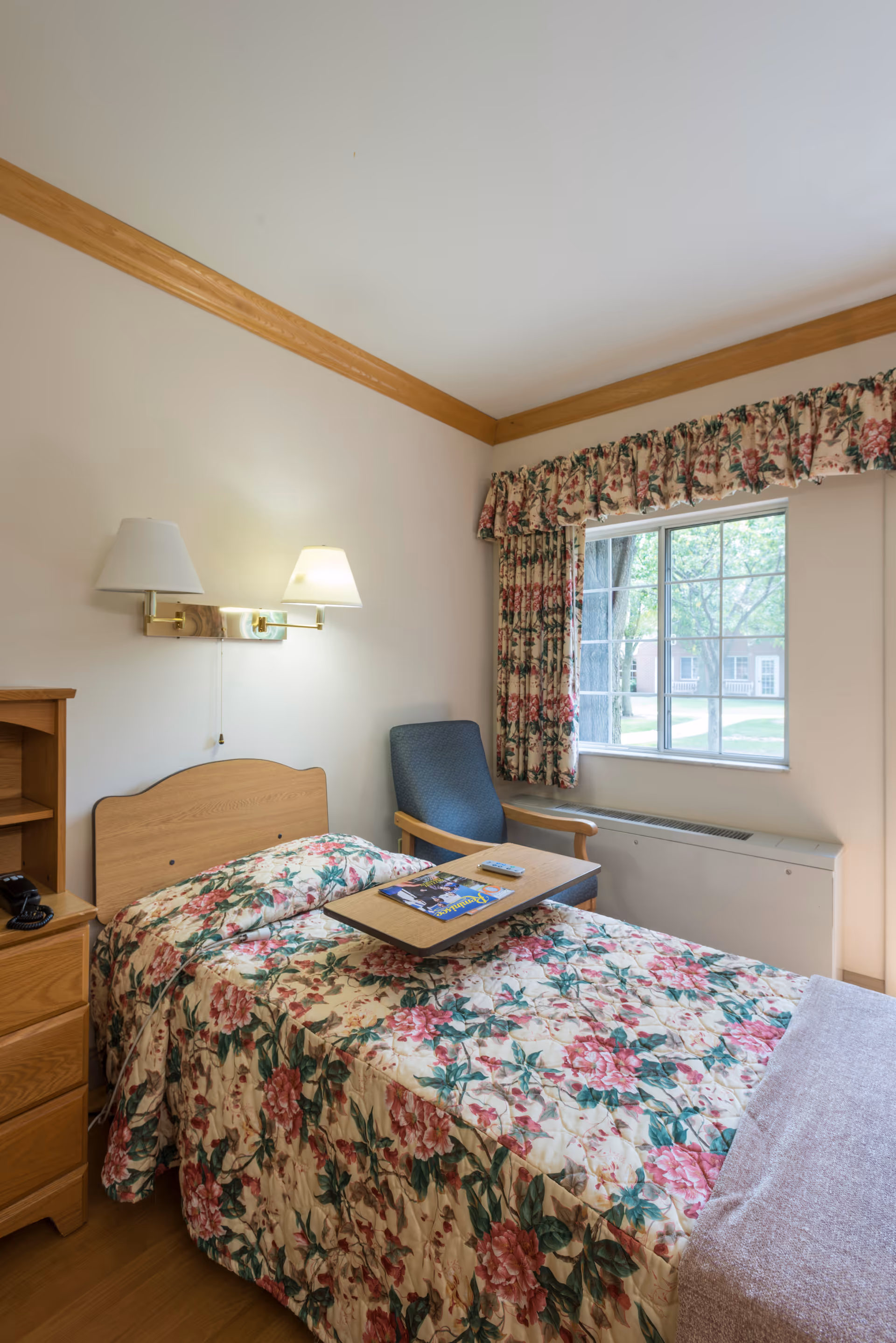 A small bedroom in a skilled nursing facility with a single bed covered in floral bedding. Next to the bed is a wooden nightstand with a telephone. Above the bed are two wall-mounted lamps. A blue armchair is positioned near a window with floral curtains, and a wooden overbed table is placed on the bed holding a magazine and a remote control. The room has light-colored walls with wooden trim and a view of trees and buildings outside the window.