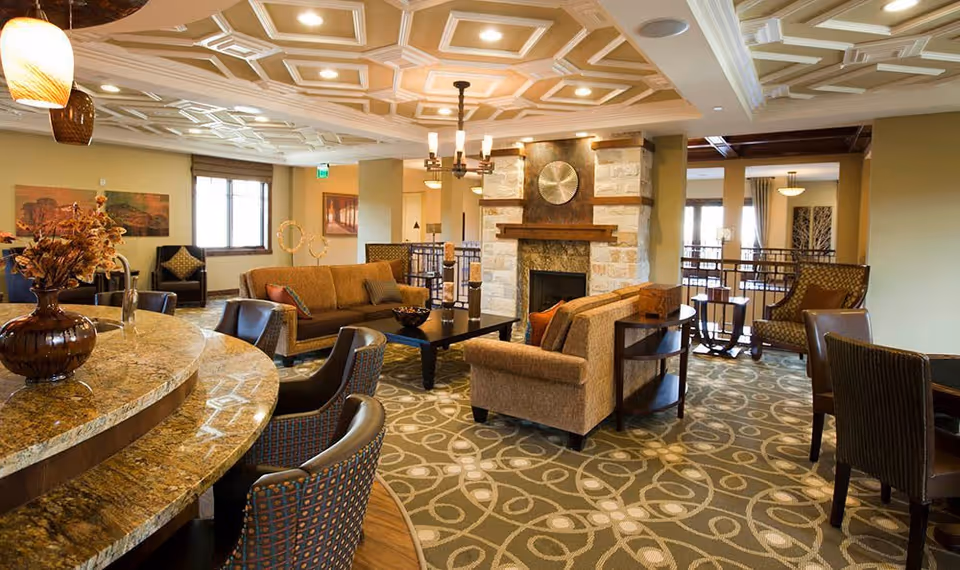 A spacious and elegantly decorated living room area in a senior living facility with comfortable sofas, armchairs, a coffee table, a stone fireplace, and a patterned carpet. The room features a coffered ceiling with recessed lighting and a chandelier, along with large windows allowing natural light to enter.