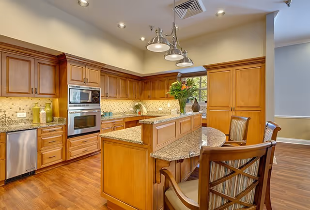 A spacious kitchen with wooden cabinets, granite countertops, and stainless steel appliances including a built-in oven and dishwasher. There is a kitchen island with a raised bar area and three wooden chairs with striped cushions. The kitchen is well-lit with recessed ceiling lights and a hanging light fixture above the island. A window with a view of greenery is visible in the background.
