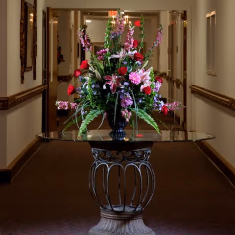 A large floral arrangement with red roses, pink lilies, and various greenery sits on a decorative glass-top table in the center of a carpeted hallway with framed artwork on the walls and an exit sign visible in the background.