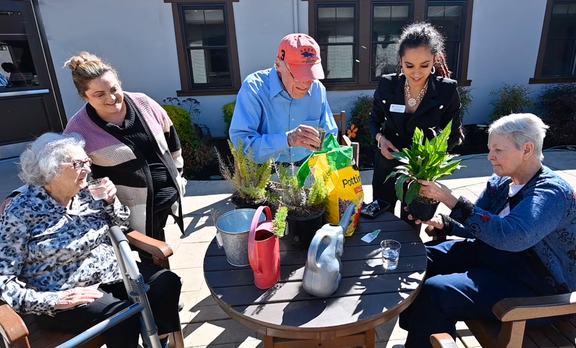 A group of five people, including elderly individuals and caregivers, are gathered around a round outdoor table with gardening supplies such as potted plants, a watering can, and potting soil. They are engaging in a gardening activity on a sunny patio outside a building with windows.