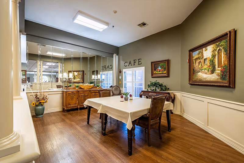Cozy cafe-style dining room with a cloth-covered rectangular table, chairs, a sideboard, mirrored wall and framed paintings.