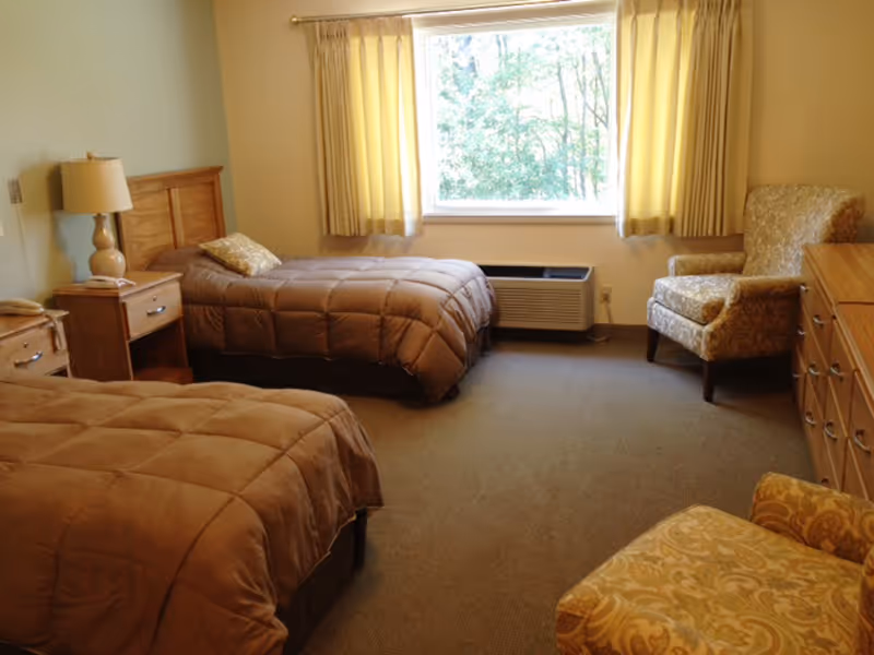A bedroom with two twin beds covered in brown comforters, a wooden nightstand with a lamp and telephone between the beds, a window with yellow curtains letting in natural light, an air conditioning unit below the window, and two upholstered armchairs with a wooden dresser on the right side.