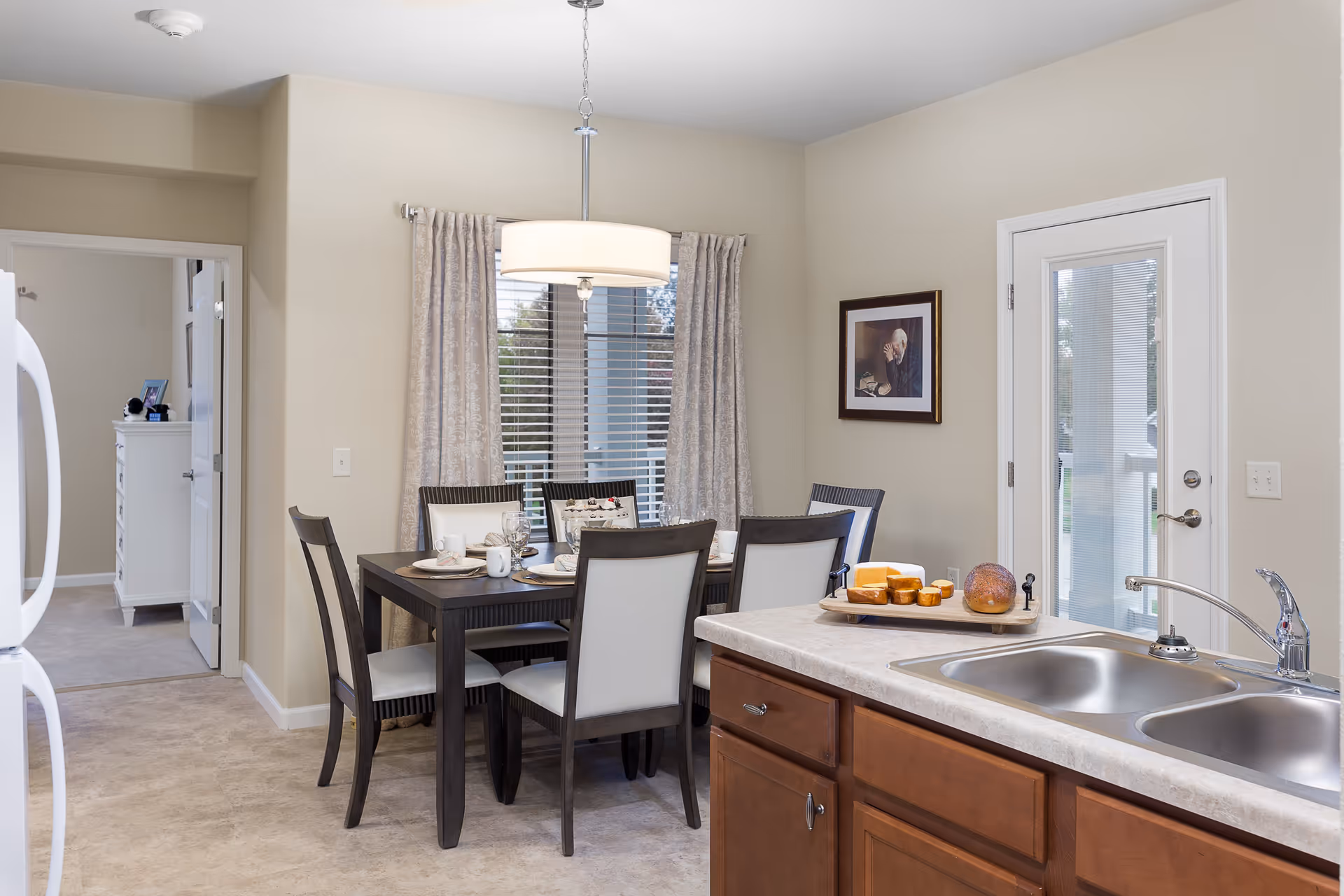 A bright dining area with a dark wooden table set for six, featuring white cushioned chairs. Behind the table is a window with beige patterned curtains and blinds. To the right, there is a kitchen counter with a double sink and a cutting board with bread and cheese. A door with glass panels is next to the counter, and a framed picture hangs on the wall. In the background, a doorway leads to another room with a white dresser.