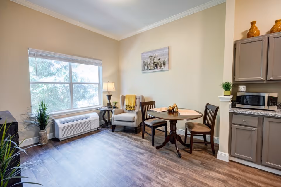 A cozy interior space with a large window letting in natural light. There is a beige armchair with a yellow throw blanket next to a small round wooden table with two chairs. A lamp and small plants are on a side table near the armchair. On the right side, there is a kitchen area with gray cabinets, a microwave, and a small plant on the counter. The floor is wood-style laminate, and a framed picture hangs on the wall above the armchair.