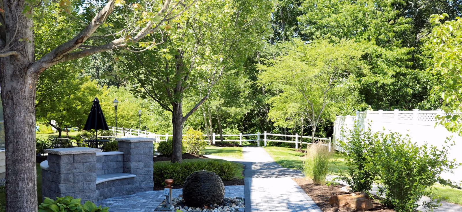 A peaceful outdoor garden area at Rockridge Retirement Community featuring a paved walkway, lush green trees and bushes, a stone bench, and a decorative stone water feature surrounded by small rocks. A white fence runs along the right side of the path.