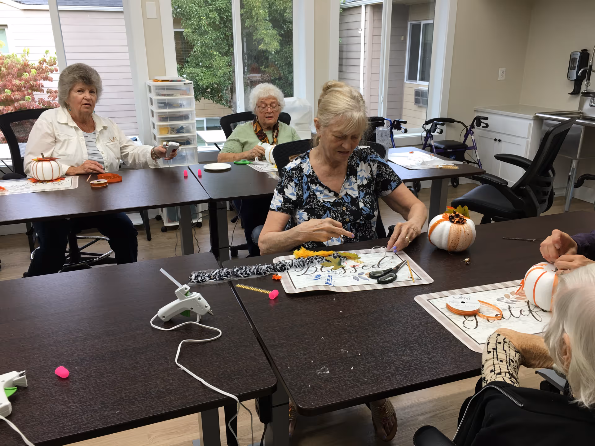 Several elderly women seated around tables in a well-lit room, engaging in a craft activity with small decorative pumpkins, ribbons, scissors, and glue guns. The room has large windows showing greenery outside and mobility aids like walkers are visible in the background.