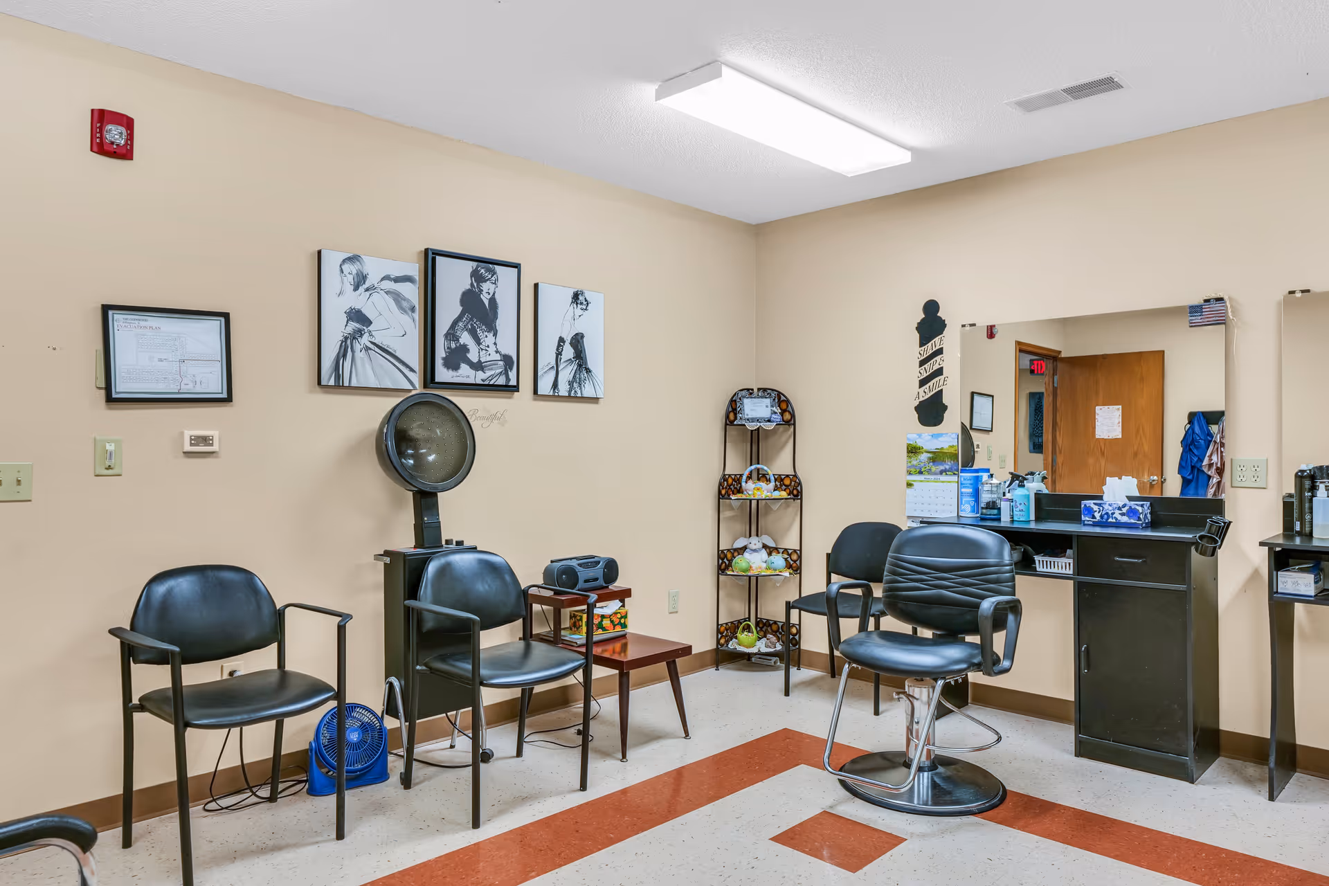 Interior of a small salon or beauty room with beige walls and a tiled floor with red stripes. There are three black chairs, including one salon chair in front of a mirror and counter with various supplies. On the wall are four black and white fashion sketches and a framed document. A small shelving unit holds decorative items, and a hair dryer hood is positioned near the chairs.