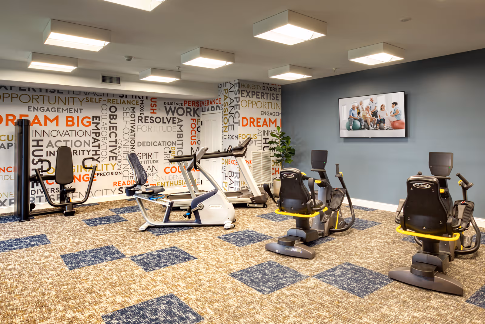 A fitness room with exercise equipment including stationary bikes, a treadmill, and a strength training machine. The room has a carpeted floor with a blue and beige pattern, a blue painted wall with a mounted TV displaying a group of elderly people exercising, and a wall covered with motivational words such as 'Dream Big', 'Work', and 'Focus'.