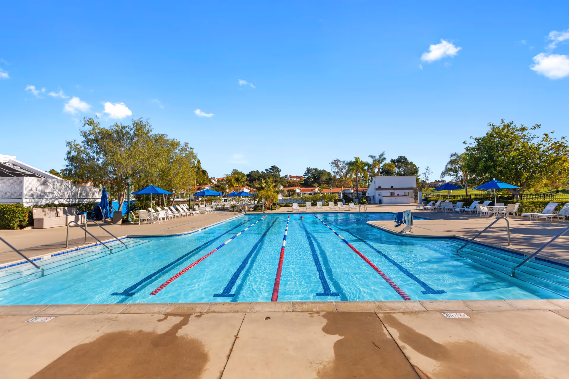 Outdoor swimming pool with lane dividers, surrounded by lounge chairs and blue umbrellas, with trees and buildings in the background under a clear blue sky.