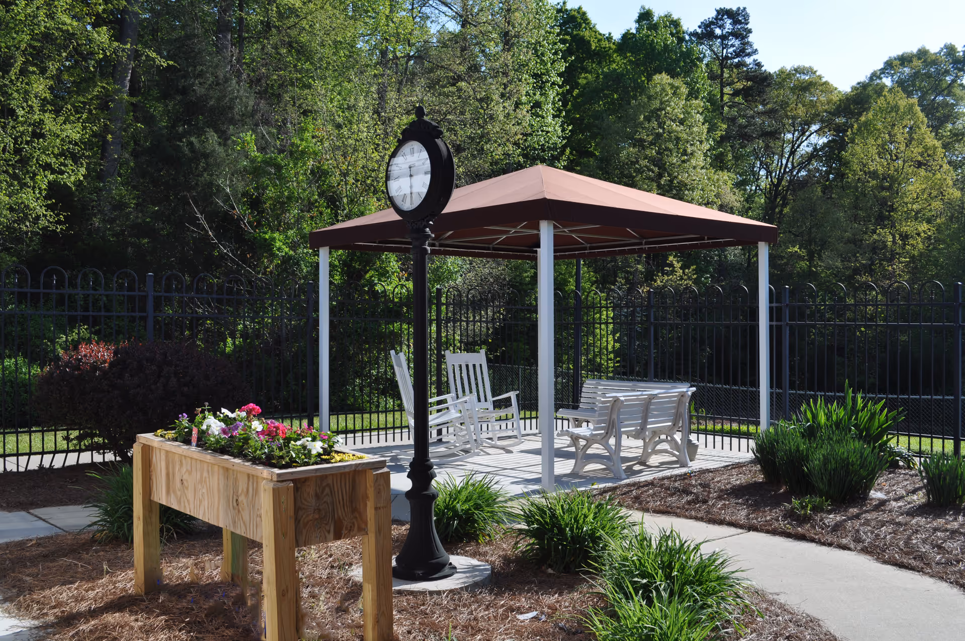 A shaded outdoor seating area with white chairs and benches under a gazebo, a freestanding clock, and a wooden planter surrounded by trees and fencing.