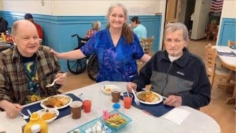 Two elderly men sitting at a round table with plates of food and drinks, accompanied by a smiling woman standing behind them with her arms around their shoulders in a dining area.