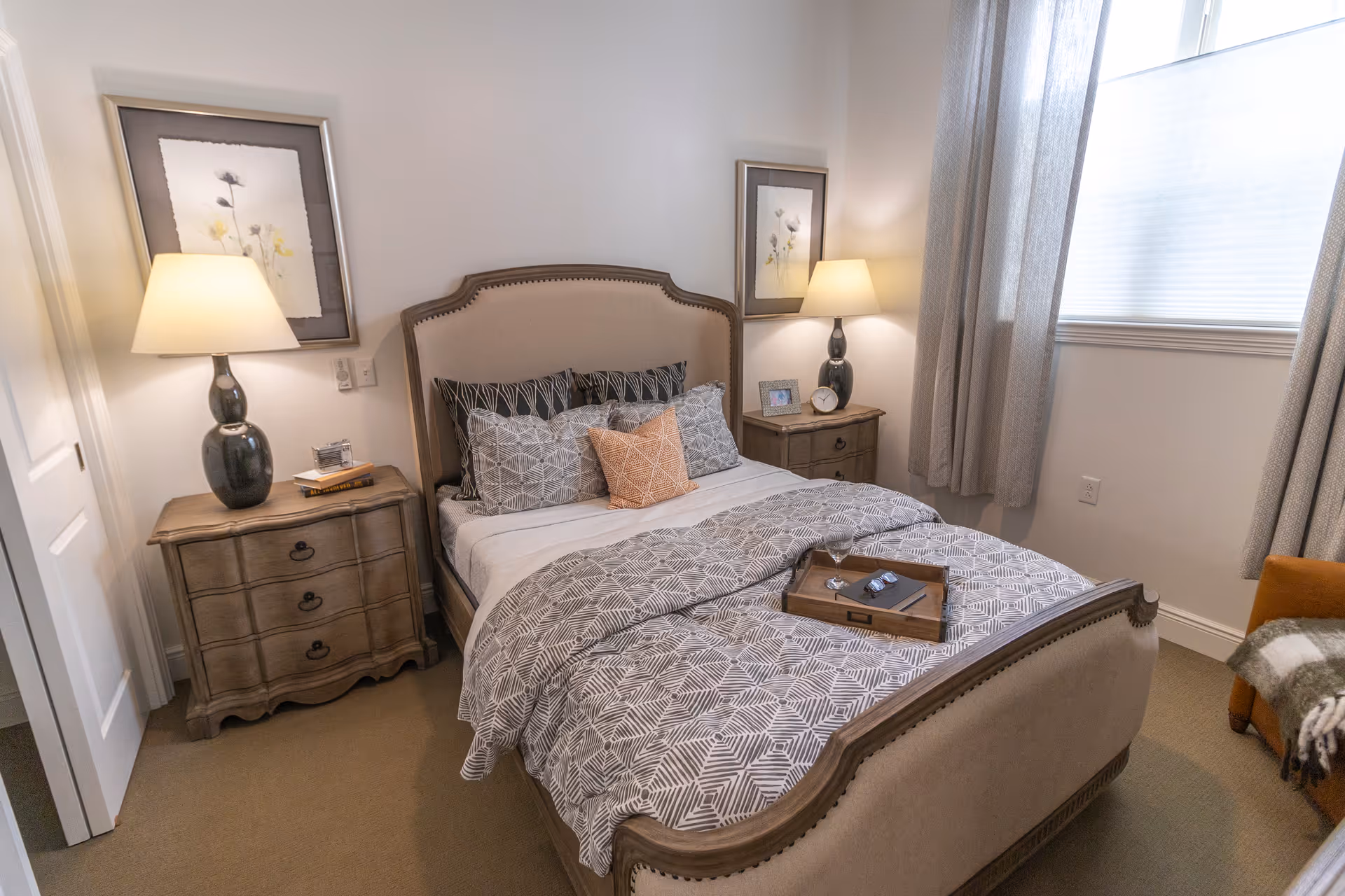 Cozy furnished bedroom with a bed dressed in patterned bedding, two bedside tables with lamps, framed wall art, and a curtained window.