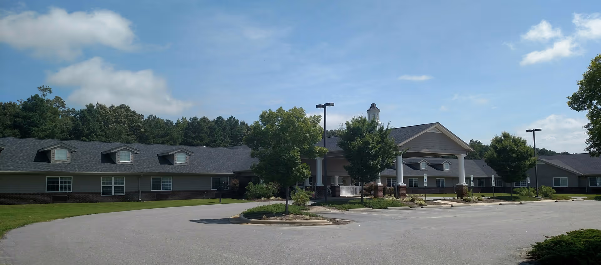 Front exterior of a single-story senior living facility with a circular driveway, columned entrance, and trees under a blue sky.