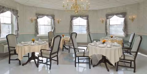 Bright dining room with round tables, chairs, and place settings under chandeliers.