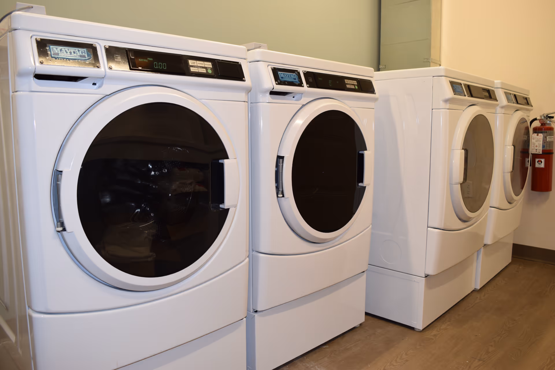 A row of four white front-loading Maytag washing machines or dryers in a laundry room with light-colored walls and wood flooring. A red fire extinguisher is mounted on the wall at the far end.