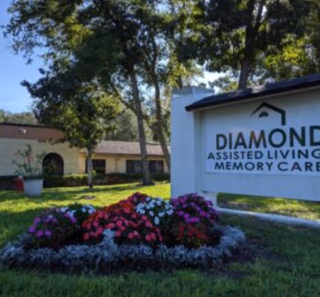 Outdoor view of Diamond Assisted Living & Memory Care facility with a white sign displaying the facility name, surrounded by a flower bed with colorful flowers and trees in the background.
