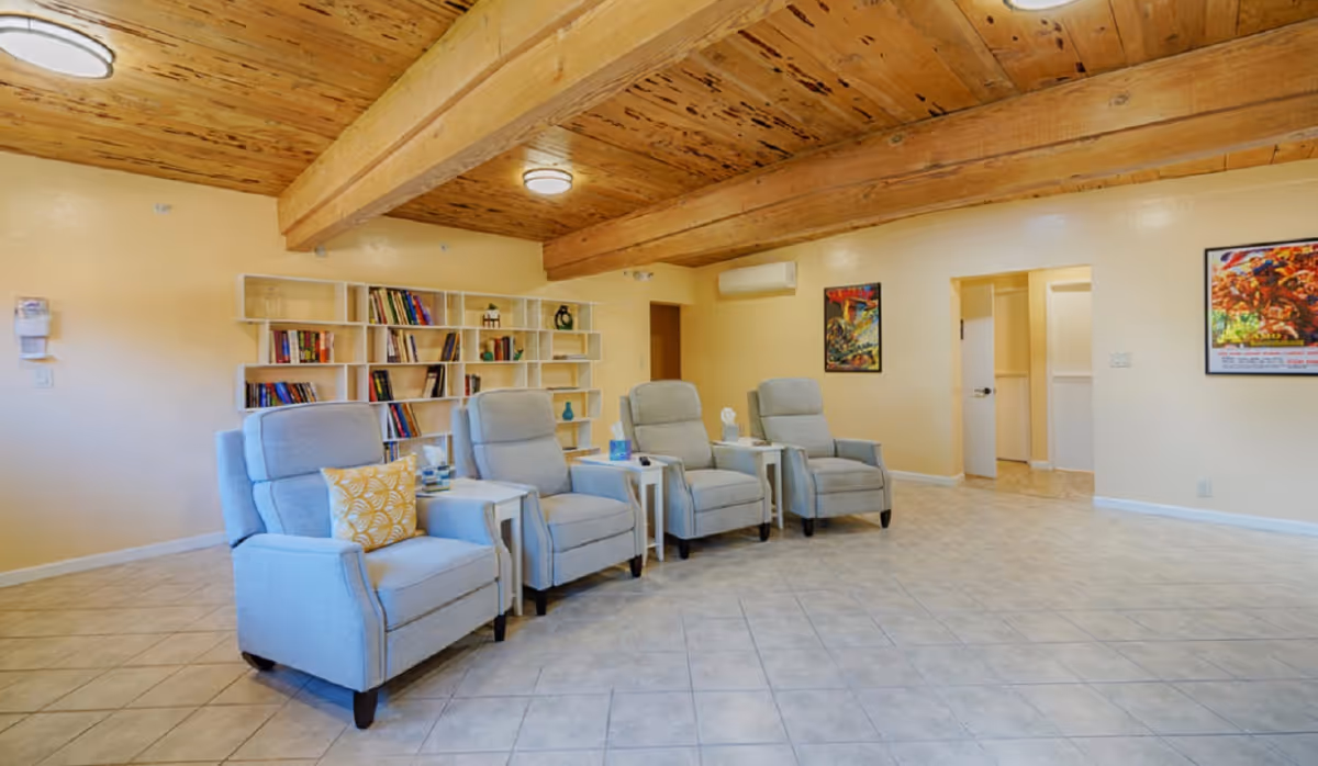Spacious seating area with four light-gray recliners, side tables, a bookshelf and exposed wooden beamed ceiling.