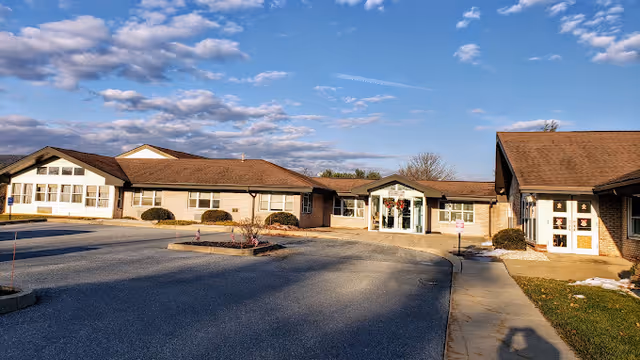 Exterior view of a single-story senior living facility building with a large parking area in front, under a partly cloudy blue sky. The building has a brown roof and light-colored walls with several windows. There is a sidewalk leading to the main entrance, which is decorated with wreaths.