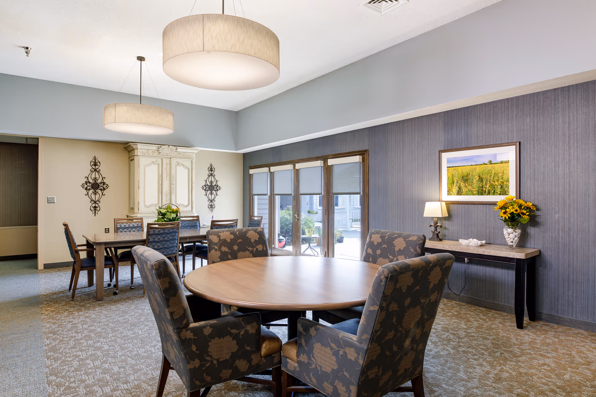 A well-lit dining room in an assisted living facility featuring a round wooden table surrounded by four upholstered chairs with floral patterns. In the background, there is a rectangular dining table with six chairs, decorative wall art, and a large cabinet. The room has large windows with blinds, a console table with a lamp and a vase of sunflowers, and two modern pendant lights hanging from the ceiling.