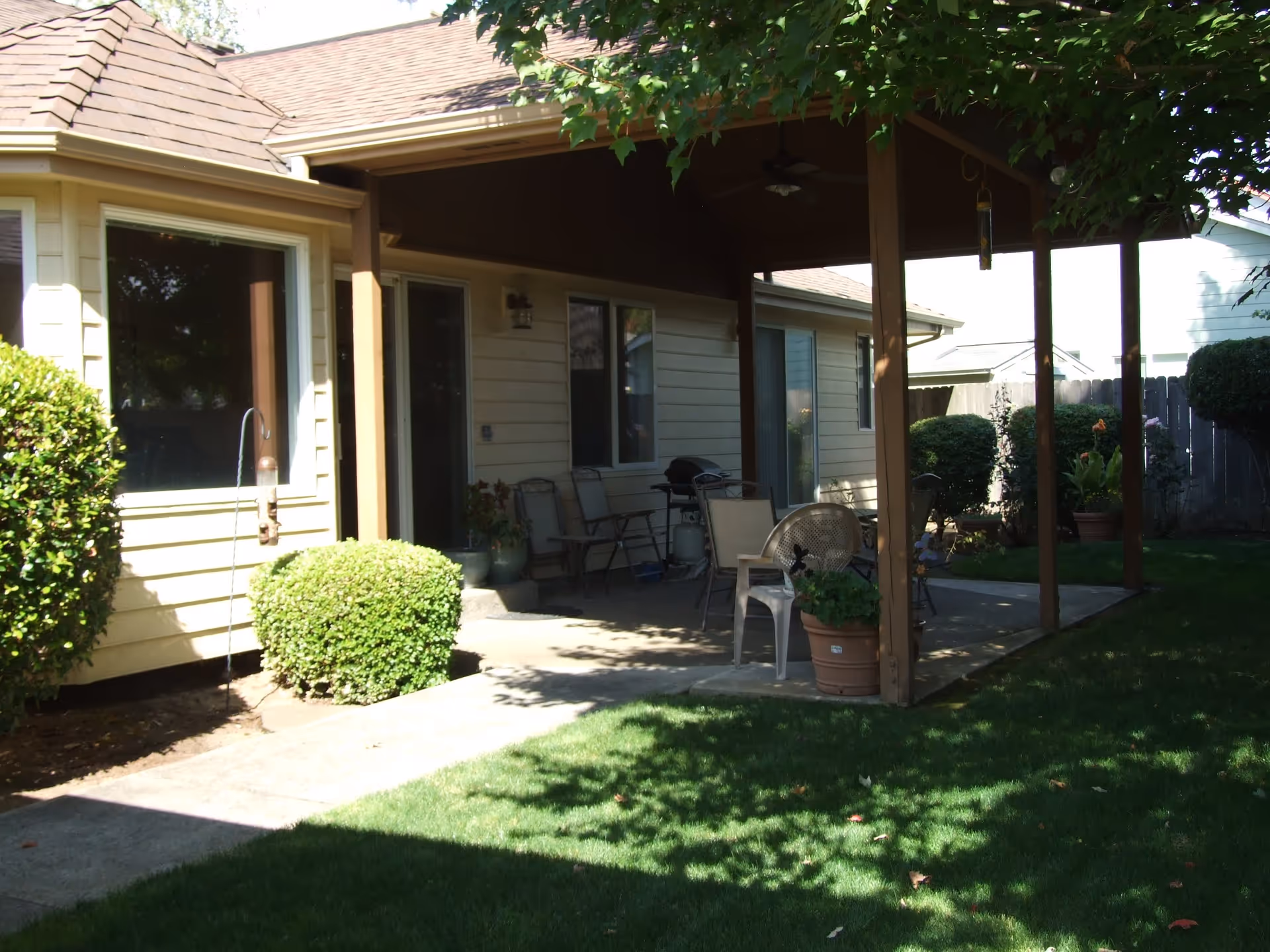 Covered patio area attached to a beige house with outdoor chairs, a table, potted plants, and a grill. The patio is surrounded by green bushes and a well-maintained lawn with trees casting shadows.