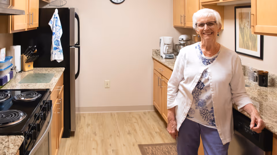 An elderly woman with white hair and glasses stands smiling in a kitchen with wooden cabinets, granite countertops, a black refrigerator, an electric stove, and various kitchen appliances. The floor is light wood, and there is a framed picture on the wall behind her.