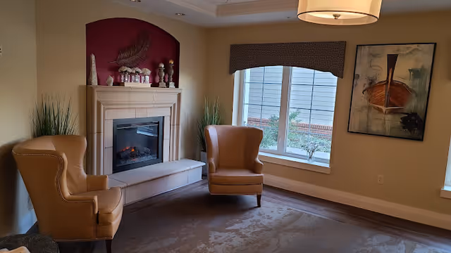 A cozy sitting area with two tan leather armchairs facing a fireplace with a white mantel. The fireplace has decorative items on top and a red accent wall behind it. There is a large window with a valance letting in natural light, and a framed abstract painting on the adjacent wall. The room has beige walls and a carpeted floor.