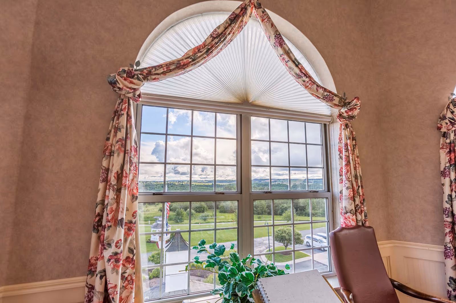View through a large window with floral curtains tied back, showing a green landscape with trees, a gazebo, and a parking area with cars. Inside, there is a brown leather chair and a table with a plant and a notebook.