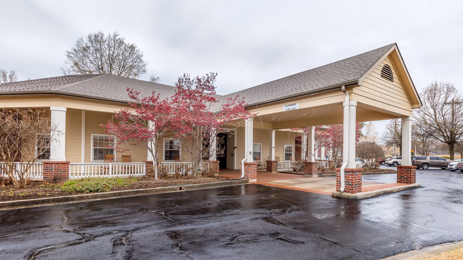 Exterior view of a single-story assisted living facility building with a covered entrance supported by white columns with brick bases. There are small trees with red leaves and some shrubs around the building. Several cars are parked in the parking lot nearby under an overcast sky.