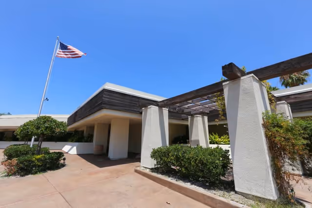 Exterior view of a single-story building with white walls and wooden accents under a clear blue sky. There is an American flag on a flagpole to the left, and a pergola structure with climbing plants in the foreground surrounded by green bushes.