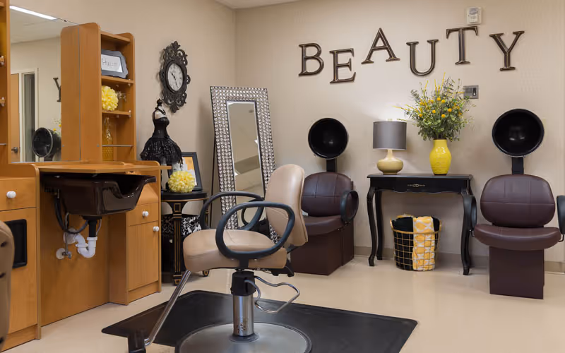 Interior of a beauty salon area in a senior living facility with a salon chair in front of a wooden vanity with a sink, two brown hair dryer chairs against the wall, a black table with a yellow vase of flowers and a lamp, and the word 'BEAUTY' displayed on the wall.