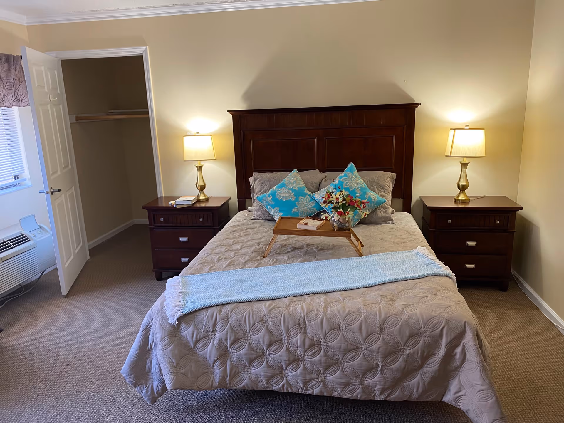 A neatly made bedroom with a bed centered between two wooden nightstands and lamps, an open closet, and a window air conditioner.