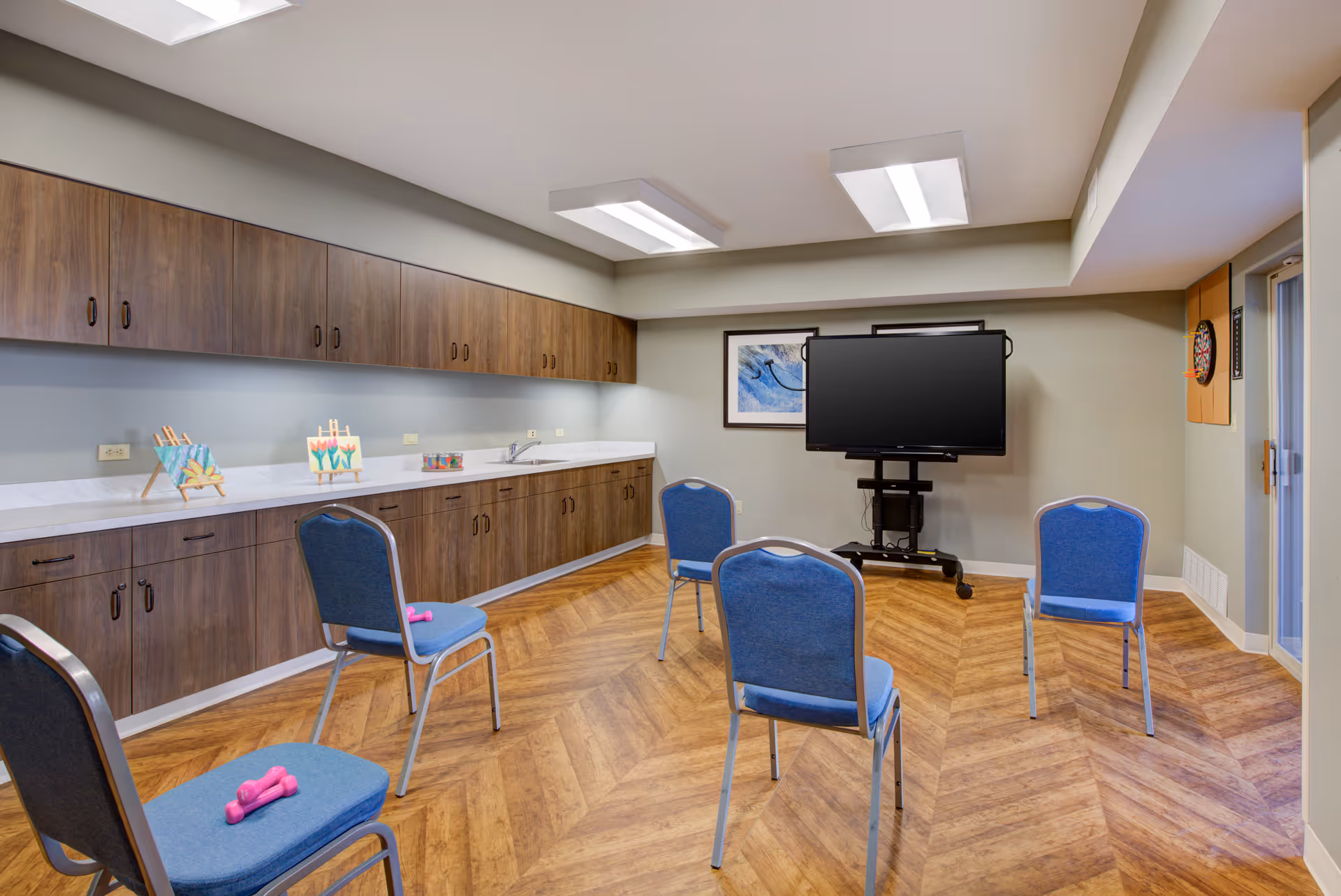 A room with wooden flooring and light gray walls featuring five blue chairs arranged in a semi-circle, each with a small pink dumbbell on the seat. There is a large flat-screen TV on a stand against the wall, a dartboard on the right wall, and wooden cabinets with a white countertop along the left wall. Two framed pictures hang on the wall behind the TV.