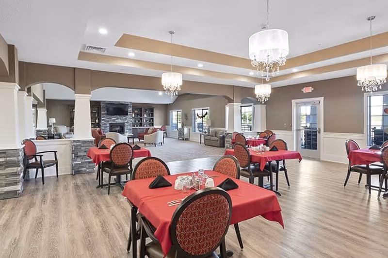 A spacious dining area in an assisted living facility with several tables covered in red tablecloths, each set with glasses, napkins, and cutlery. The room features wooden flooring, beige walls with white wainscoting, and elegant chandeliers hanging from the ceiling. In the background, there is a living area with sofas and a stone fireplace with built-in shelves.