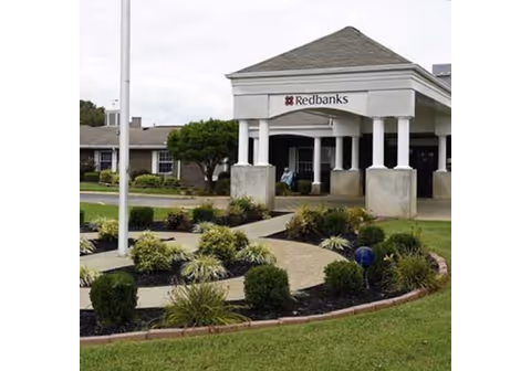 Entrance of the Redbanks facility featuring a covered driveway with white pillars, a landscaped garden with bushes and plants, and a flagpole on a grassy lawn.
