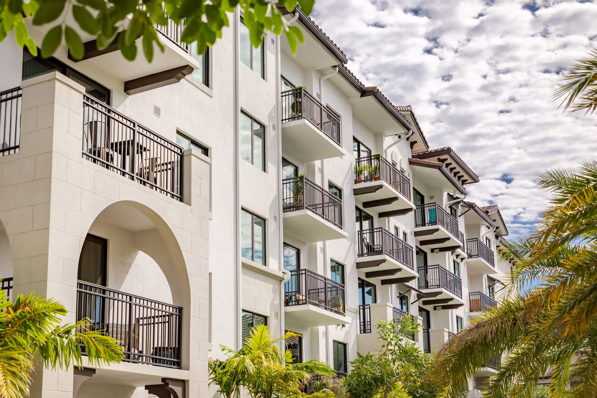 Exterior view of a multi-story residential building with white walls, multiple balconies with black railings, and tropical palm trees in the foreground under a partly cloudy sky.