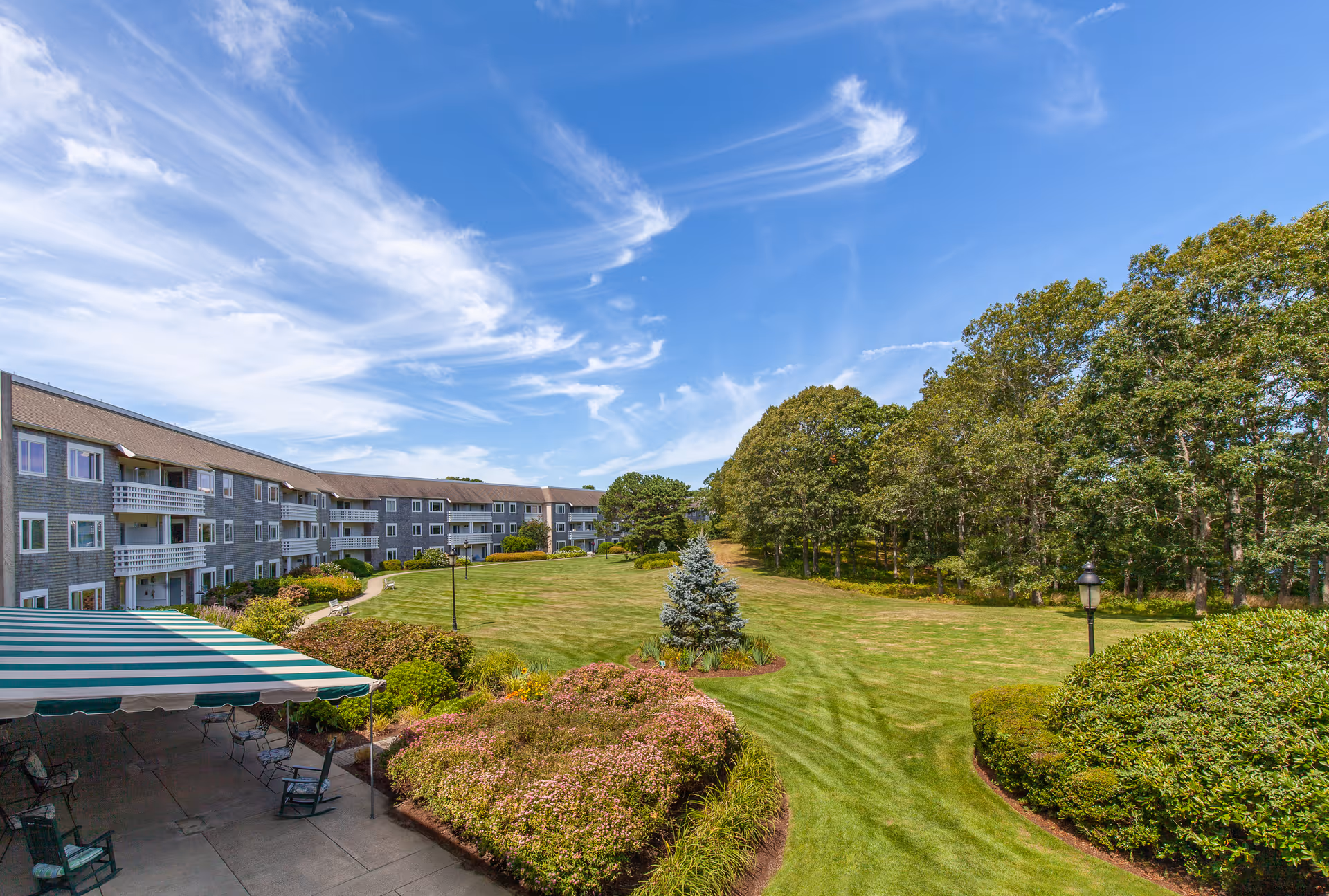 Outdoor view of Thirwood Place showing a large green lawn with manicured bushes and trees, a multi-story residential building with balconies on the left, and a patio area with striped green and white awning and chairs underneath. The sky is blue with wispy clouds.