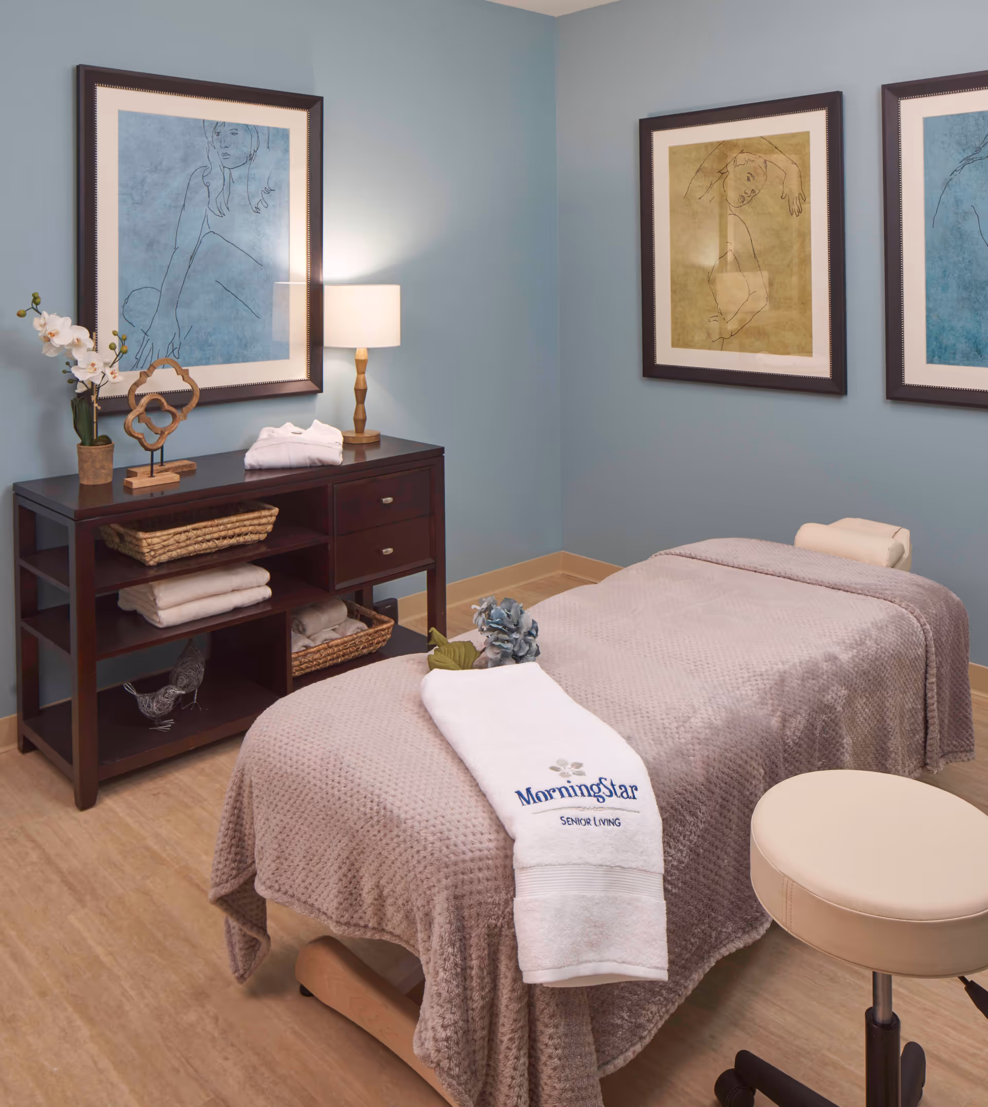 A serene massage or therapy room with a massage table covered in a textured beige blanket and a white towel embroidered with 'MorningStar Senior Living'. The room has light blue walls adorned with three framed minimalist line art pictures. A dark wooden cabinet with shelves and drawers holds neatly folded towels, decorative items, and a table lamp emitting a warm light. A beige cushioned stool is positioned near the massage table.