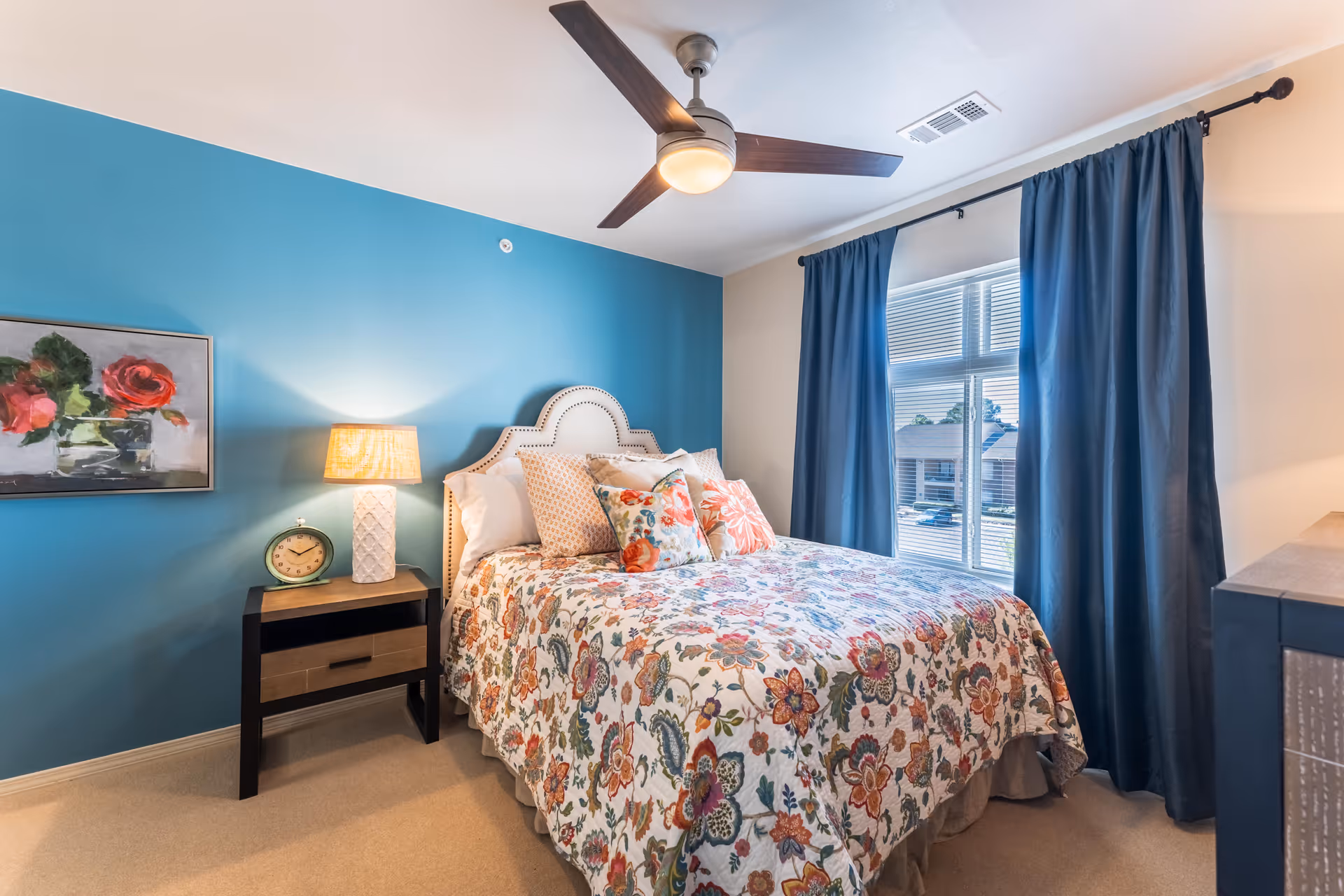 A cozy bedroom with a floral patterned bedspread on a bed with a white upholstered headboard. There is a nightstand with a lamp and a clock on it next to the bed. The wall behind the bed is painted blue and has a framed painting of flowers. A window with blue curtains allows natural light into the room, and a ceiling fan with a light fixture is mounted on the ceiling.