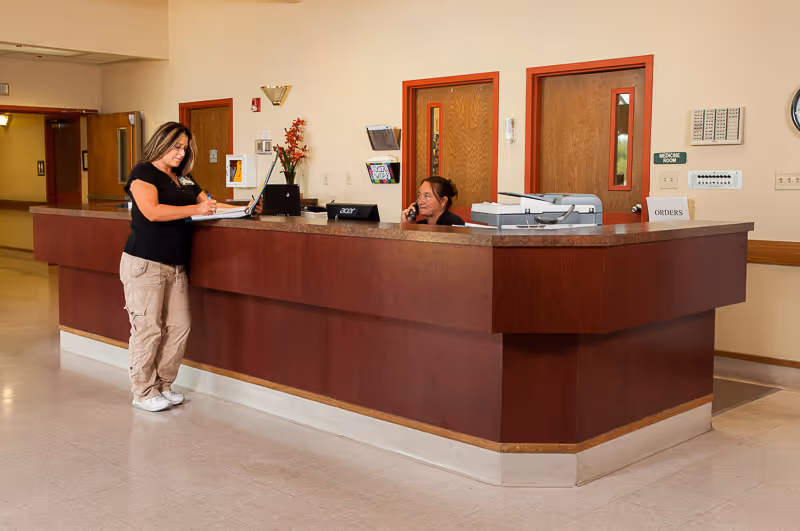 A reception area inside a senior living facility with a wooden front desk. One woman is standing and writing on a clipboard, while another woman is sitting behind the desk talking on the phone. There are doors and a sign labeled 'Medicine Room' on the wall behind the desk.