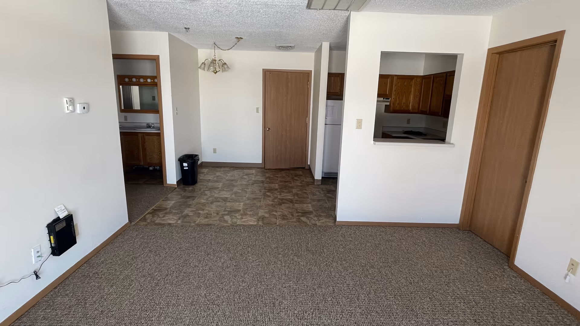 Empty interior space showing a small dining area with tiled flooring, a kitchen with wooden cabinets visible through a pass-through window, and a carpeted living area. There are two closed wooden doors and an open doorway leading to a bathroom with a sink and mirror. A black trash can is placed near the dining area.