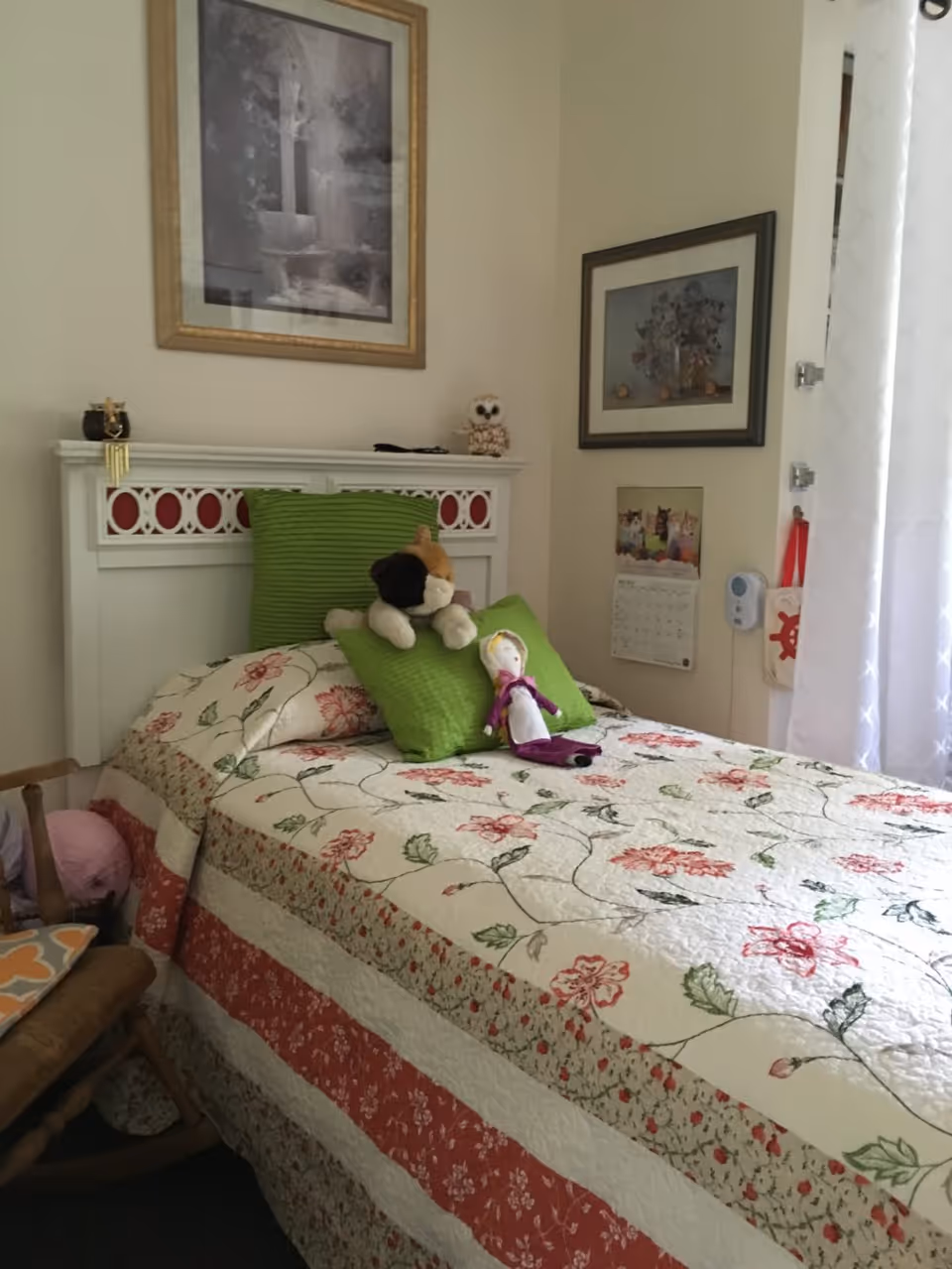 A cozy bedroom with a single bed covered in a floral quilt and two green pillows. On the bed are a stuffed dog toy and a doll. The headboard is white with red decorative insets. Above the bed are two framed pictures on the wall. To the right is a window with white curtains, a wall calendar featuring cats, and a small hanging bag with a red design. A wooden rocking chair with a cushion is partially visible on the left side.