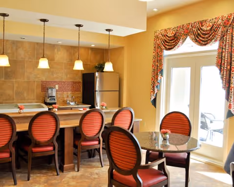A cozy dining area with a kitchen counter featuring four red cushioned chairs. The kitchen area has a refrigerator, coffee maker, and a tiled backsplash. There is a round table with four matching red cushioned chairs near a glass door with floral curtains, allowing natural light to enter the room.