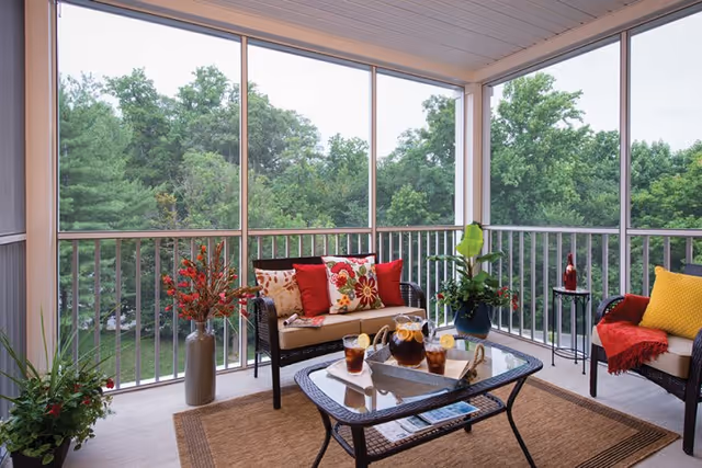 A screened-in porch with a view of green trees outside. The porch has a wicker loveseat with colorful floral and red cushions, a glass-top coffee table with a pitcher of iced tea and glasses, and a wicker chair with a yellow cushion and red throw blanket. There are several potted plants and a small side table with a decorative bottle.