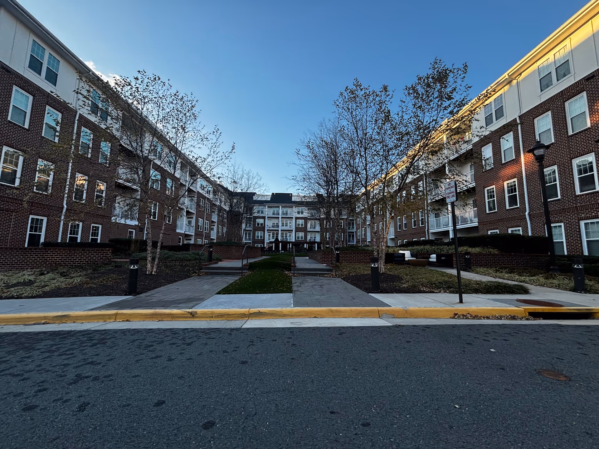 Brick multi-story senior living building forming a U-shaped courtyard with trees and walkways under a clear sky.