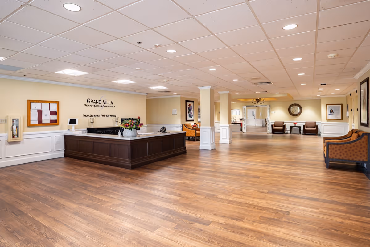 Spacious reception area of Grand Villa Senior Living Community with a large wooden front desk, flower arrangement, seating areas with orange patterned chairs, wall clock, framed pictures, and wood flooring under a white tiled ceiling with recessed lighting.