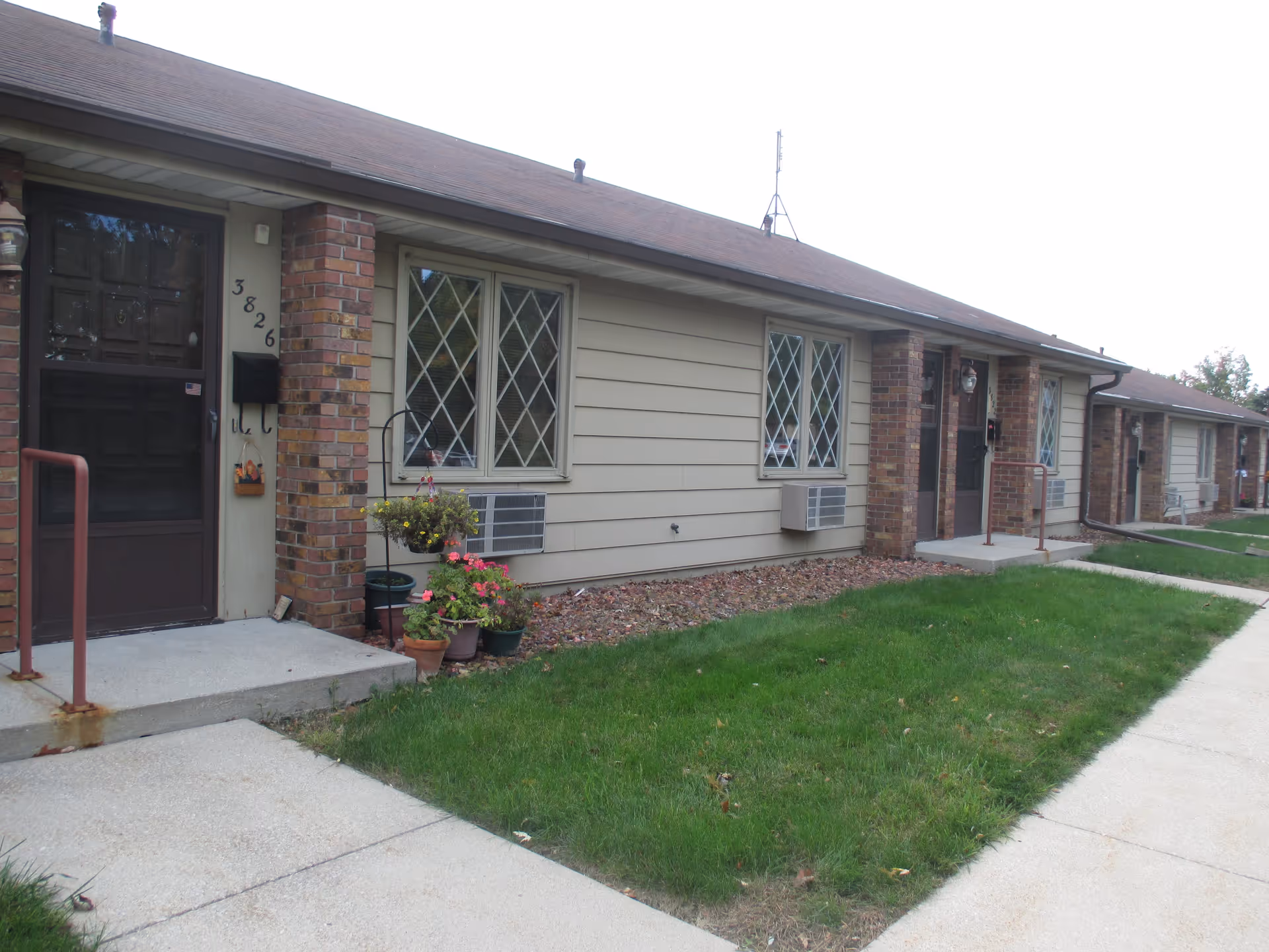 Row of single-story apartment entrances with brick columns, diamond-pane windows, window air conditioners, potted plants, and a concrete walkway beside a small lawn.