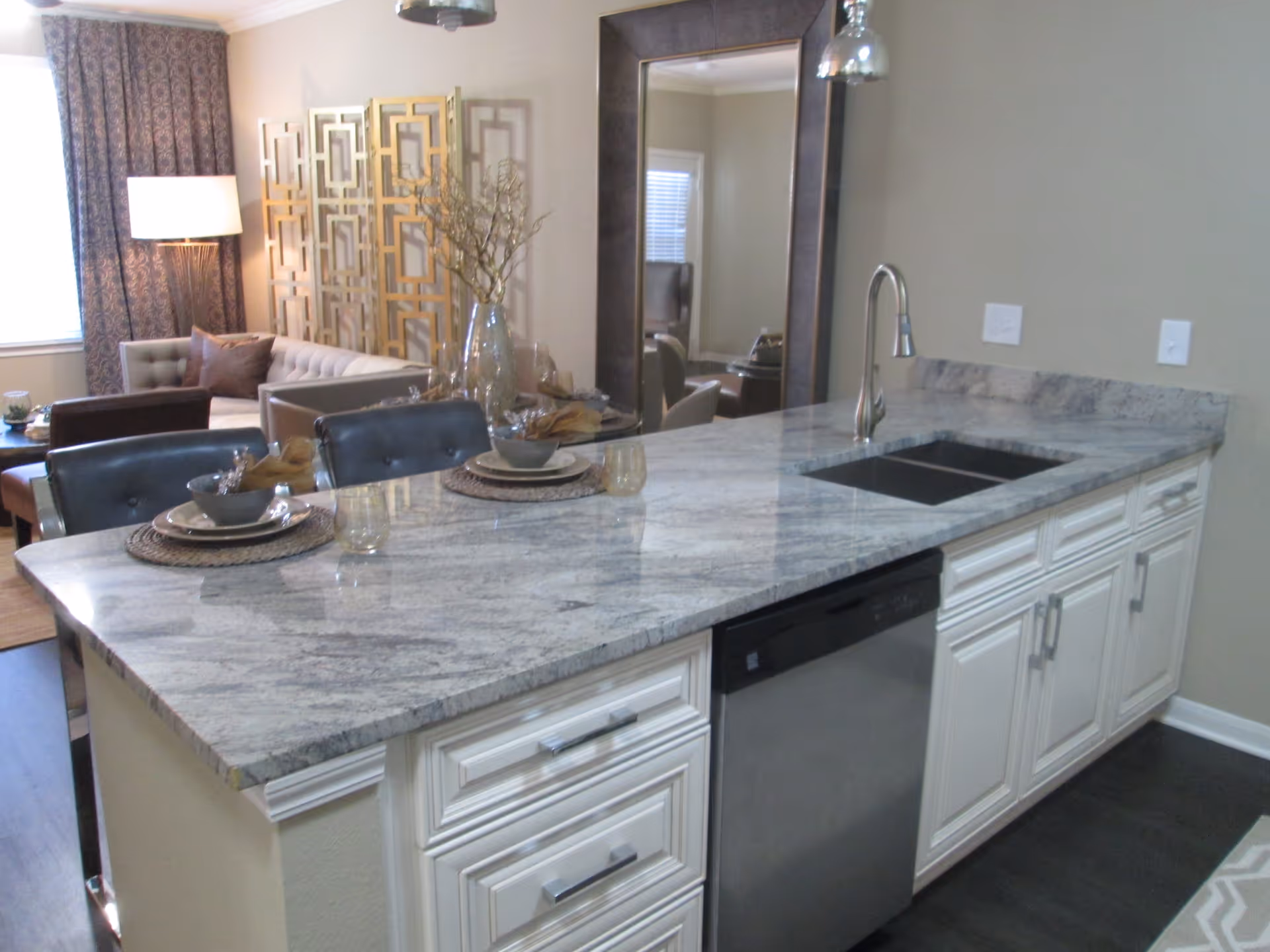 Modern kitchen island with a gray marble countertop, a built-in sink, and a stainless steel dishwasher. Two place settings with bowls, plates, and glasses are arranged on the island. In the background, there is a living room area with a beige tufted sofa, a floor lamp, decorative room divider, and a large mirror on the wall.